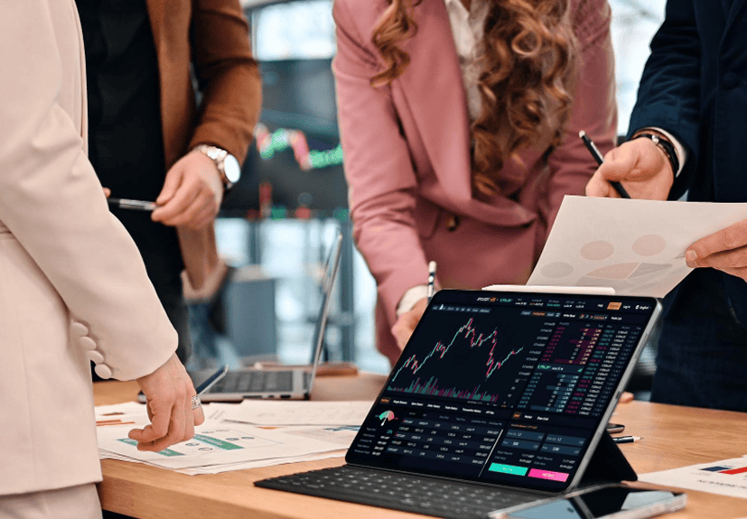 Four people in business attire review financial charts and documents at a desk, with a laptop displaying stock market data and graphs during interactive Live Sessions.