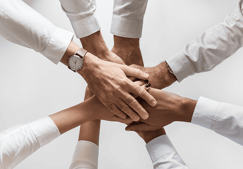 Five people stack their hands together in the center, symbolizing teamwork or unity, against a neutral background.