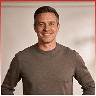 A man with short brown hair, wearing a brown sweater, stands and smiles in front of a plain light-colored background.