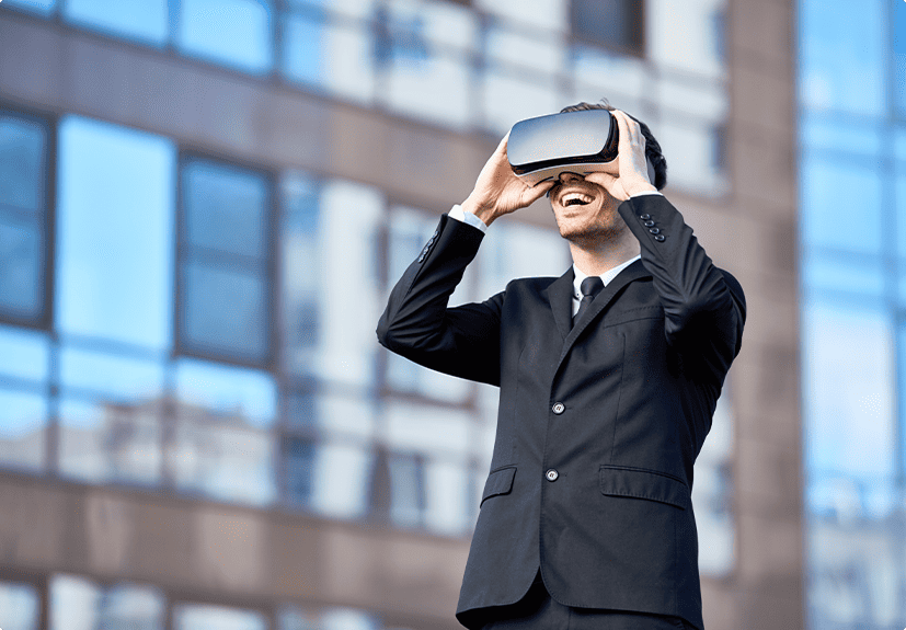 A man in a suit stands outside a glass building, smiling while wearing a virtual reality headset.