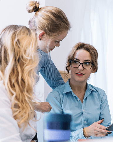 Three women in business attire are having a discussion in an office setting, with one woman sitting and the other two standing nearby.