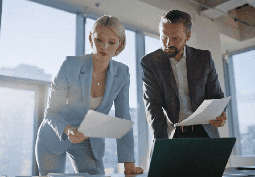 Two business professionals review documents together at a desk in a modern office with large windows and natural light.