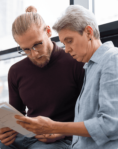 A younger man with glasses and an older woman are sitting together, looking at documents or papers in their hands, indoors by a window.