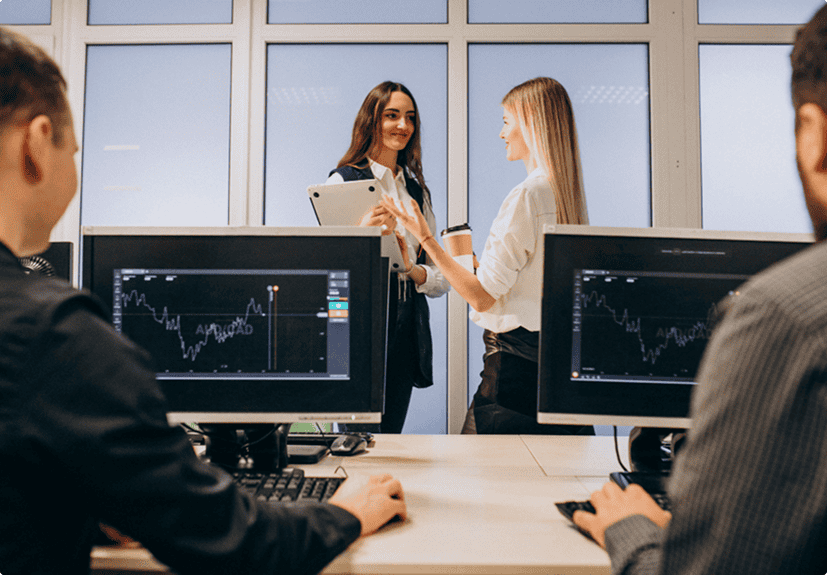 Two women standing and talking near windows, one holding a tablet, while two people sit at computers displaying financial graphs in the foreground.