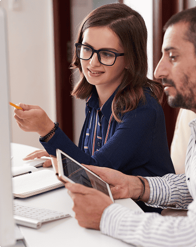 A woman points at a computer monitor while speaking to a man holding a tablet; both are seated at a desk with a keyboard and notebook.