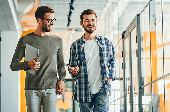 Two men walking indoors, one holding a laptop, both engaged in conversation and smiling in a modern office environment.