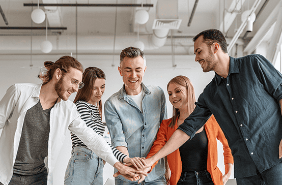 Five people stand in a circle indoors, placing their hands together in the center, suggesting teamwork or collaboration in an office setting.