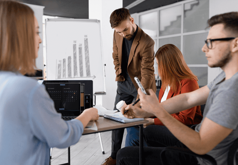 Four people in an office setting have a meeting; one man stands near a whiteboard with a bar graph, while the others sit at a table equipped for Live Sessions with a laptop, notebook, and phone.