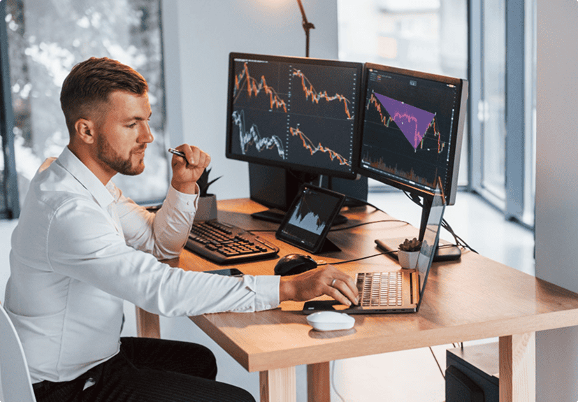 A man in business attire sits at a desk analyzing stock trading charts on multiple computer monitors in a modern office setting.