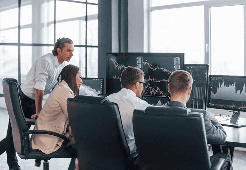 Four people in an office look at computer monitors displaying financial charts and graphs, suggesting analysis or trading activity.
