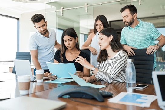 Five people sit and stand around a conference table with laptops, papers, and coffee cups, discussing documents in an office meeting room.
