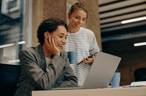 Two people in an office setting look at a laptop screen together, one sitting and smiling while the other stands holding a mug and tablet.